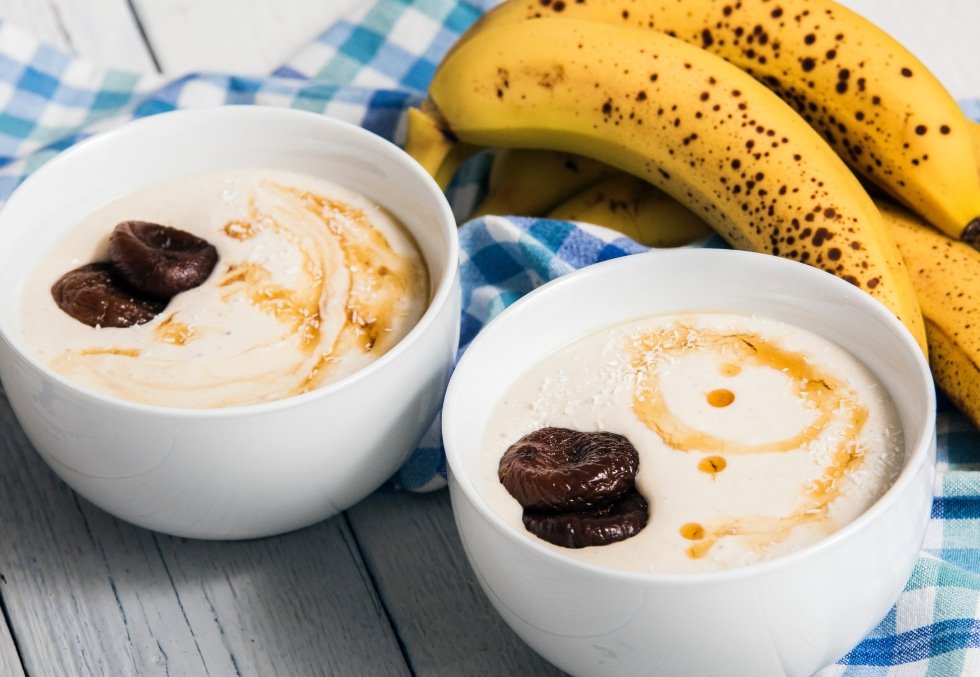 Two bowls of yogurt topped with figs and honey, with ripe bananas and a blue plaid cloth in the background.